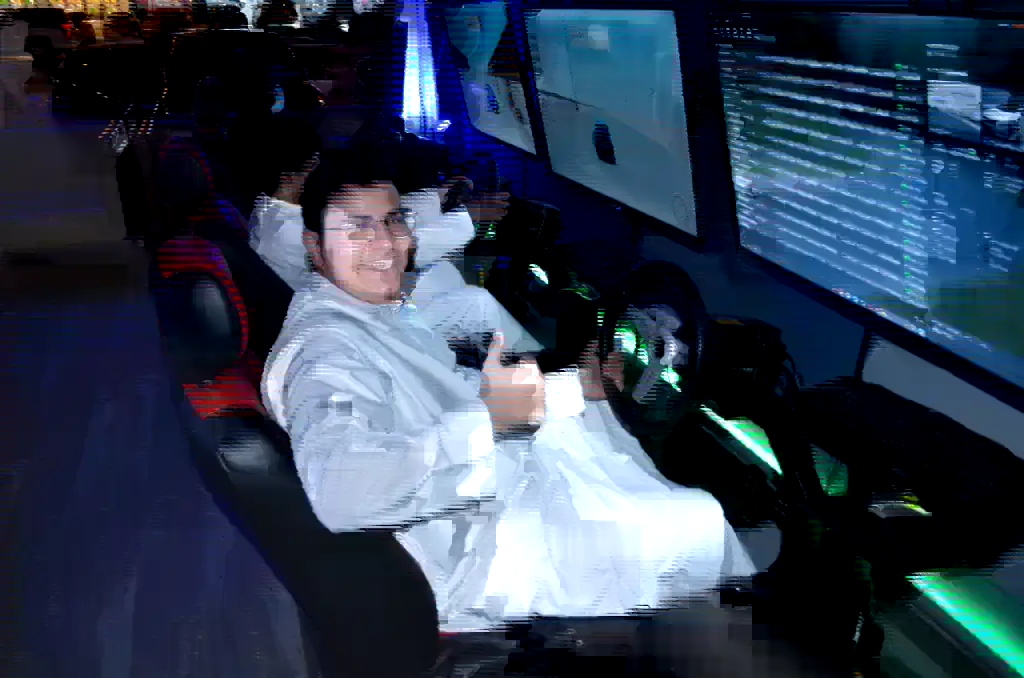 شاب سعودي بثوب أبيض جالس على مقعد سباق أحمر وأسود داخل أسيتو كلوب، ممسكًا الدركسون أمام ثلاث شاشات تعرض لعبة قيادة، وإضاءة خضراء تحت الجهاز